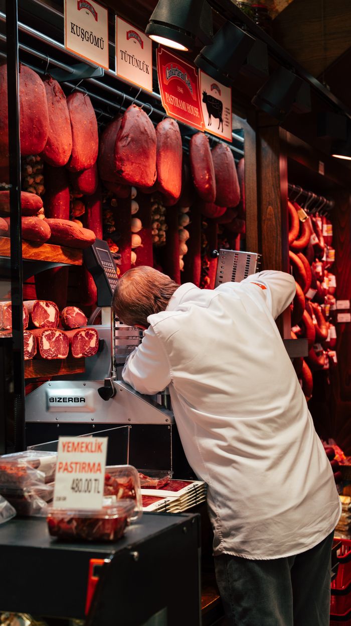 A butcher in Istanbul prepares meat surrounded by hanging cured meats and sausages.